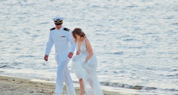 A military man in uniform walking on the beach with woman in wedding dress. 
