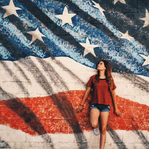 A young adult woman standing against concrete wall decorated with American flag.