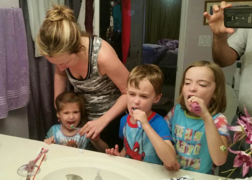 Mom helping 3 kids brush teeth in bathroom.